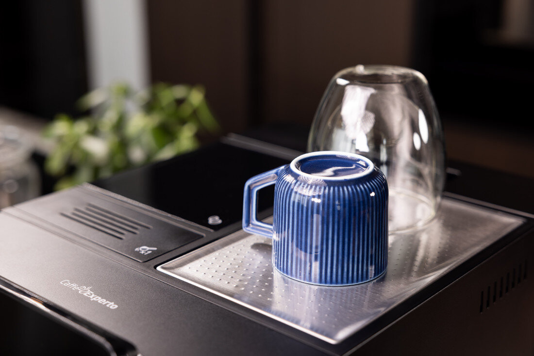 Na górnej części ekspresu do kawy znajdują się odwrócona niebieska filiżanka i szklanka. Na urządzeniu widoczny jest napis \'CaffeExperto\'.