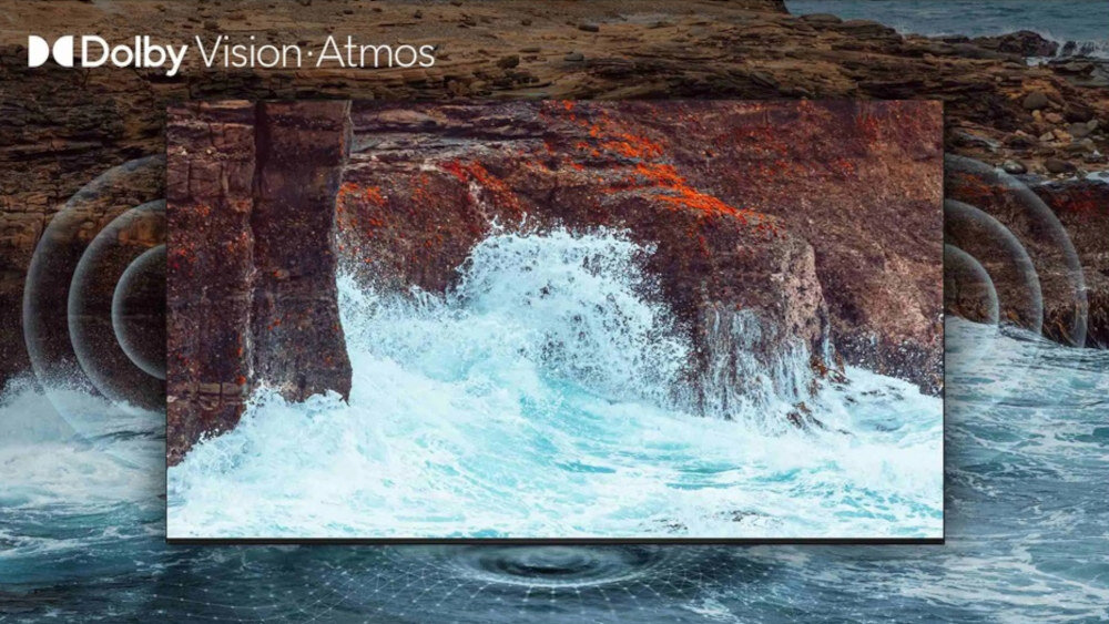 Obraz przedstawia wyświetlacz telewizora z falami uderzającymi o skały, z efektem dźwięku przestrzennego. W lewym górnym rogu widnieje napis \'Dolby Vision Atmos\'.