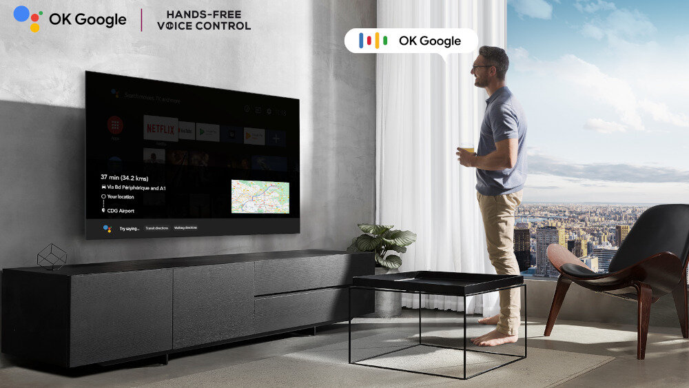 W salonie na ścianie wisi duży telewizor z wyświetlonym interfejsem aplikacji, obok stoi mężczyzna. Na górze obrazu widnieje napis \'OK Google HANDS-FREE VOICE CONTROL\'.