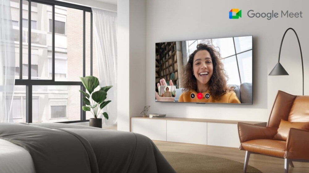 W nowoczesnym salonie na ścianie wisi duży telewizor, na którym wyświetlana jest wideorozmowa z uśmiechniętą osobą. W prawym górnym rogu ekranu widnieje logo Google Meet.