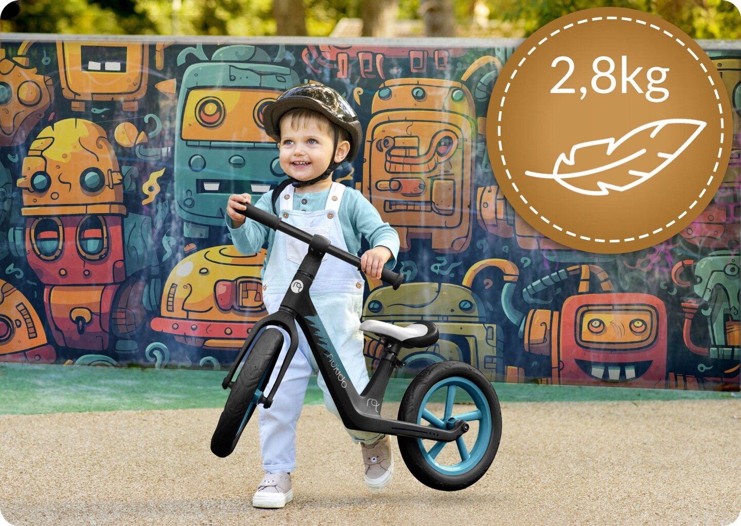 Dziecko w kasku trzyma czarny rowerek biegowy na tle kolorowej ściany z rysunkami robotów. W prawym górnym rogu widoczna jest ikona piórka z napisem \'2,8kg\'.