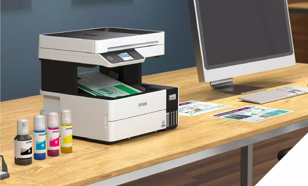 Na biurku znajduje się urządzenie wielofunkcyjne z widocznym logo EPSON, obok którego stoją cztery butelki z tuszem w różnych kolorach. Obok urządzenia znajduje się monitor, klawiatura i kilka kolorowych wydruków.