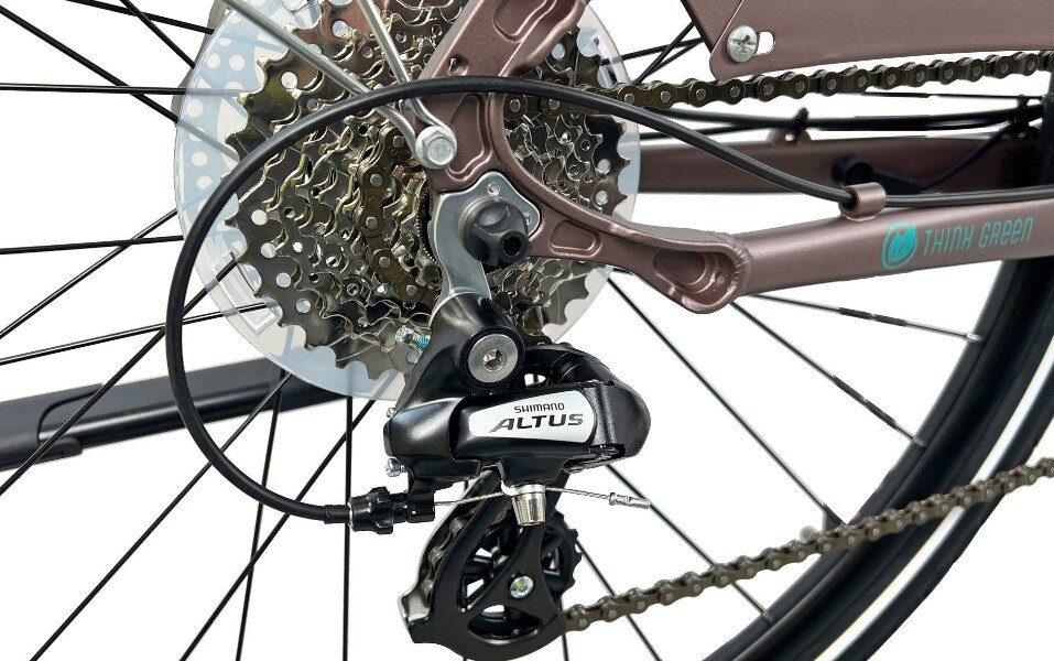 Zbliżenie na mechanizm przerzutki rowerowej z napisem \'SHIMANO ALTUS\' oraz fragment tylnego koła z zębatkami. Na ramie roweru widoczny jest napis \'THINK GREEN\'.