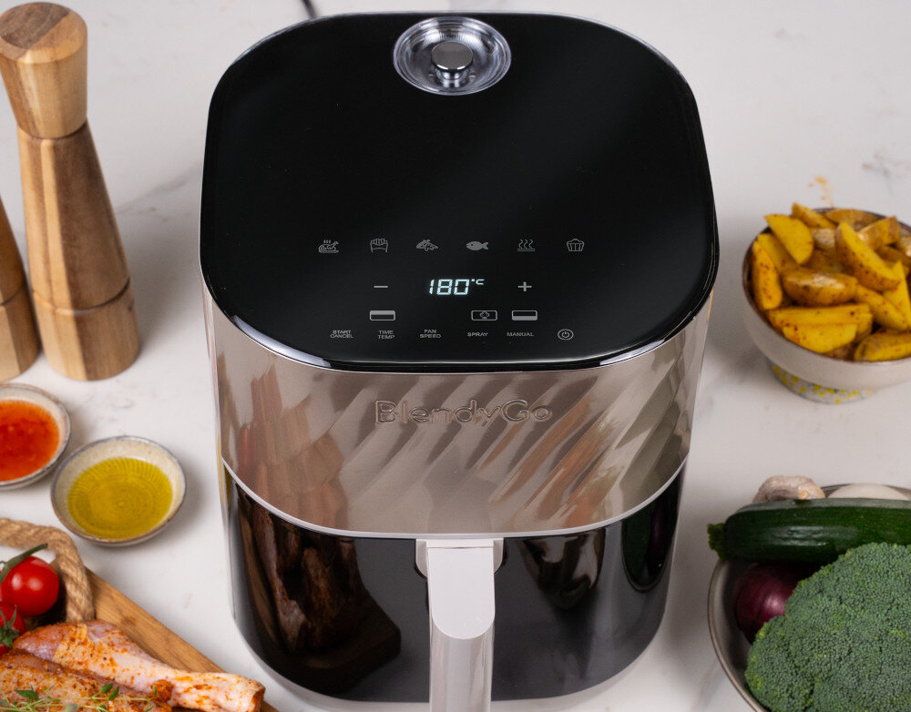 Na obrazie widoczna jest frytkownica beztłuszczowa BlendyGo z wyświetlaczem pokazującym temperaturę 180°C. Wokół urządzenia znajdują się różne składniki, takie jak przyprawy, warzywa i kawałki mięsa.