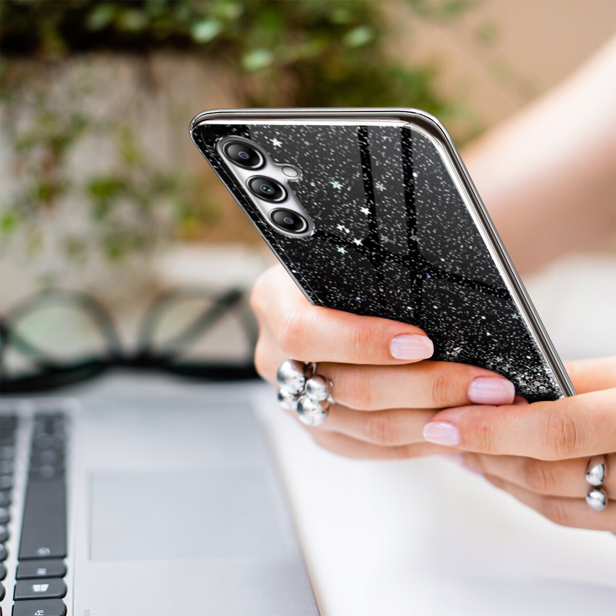 Osoba trzyma telefon z czarnym etui pokrytym brokatem i małymi gwiazdkami. W tle widoczna jest klawiatura laptopa i okulary.