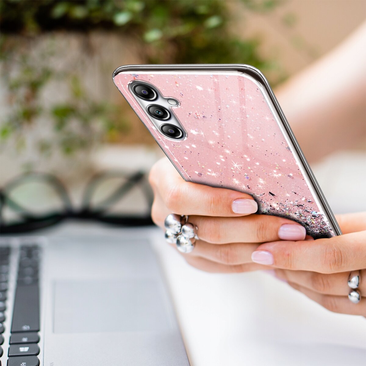 Osoba trzyma telefon z różowym, brokatowym etui, na którym widoczne są trzy obiektywy aparatu. W tle znajduje się klawiatura laptopa i okulary.