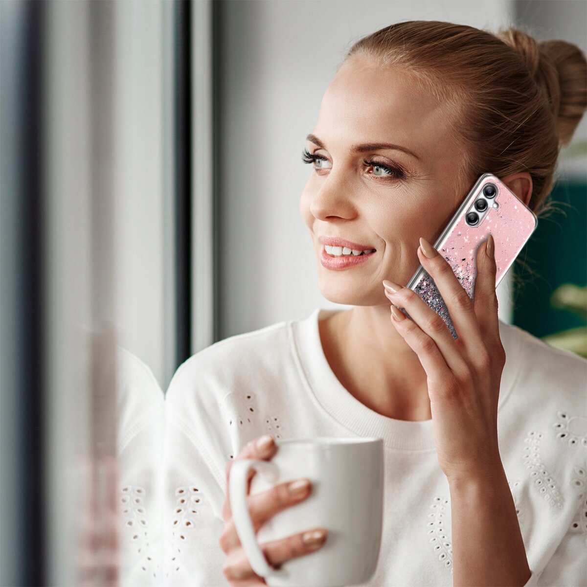 Osoba trzyma telefon z różowym, brokatowym etui przy uchu. W drugiej ręce trzyma biały kubek.