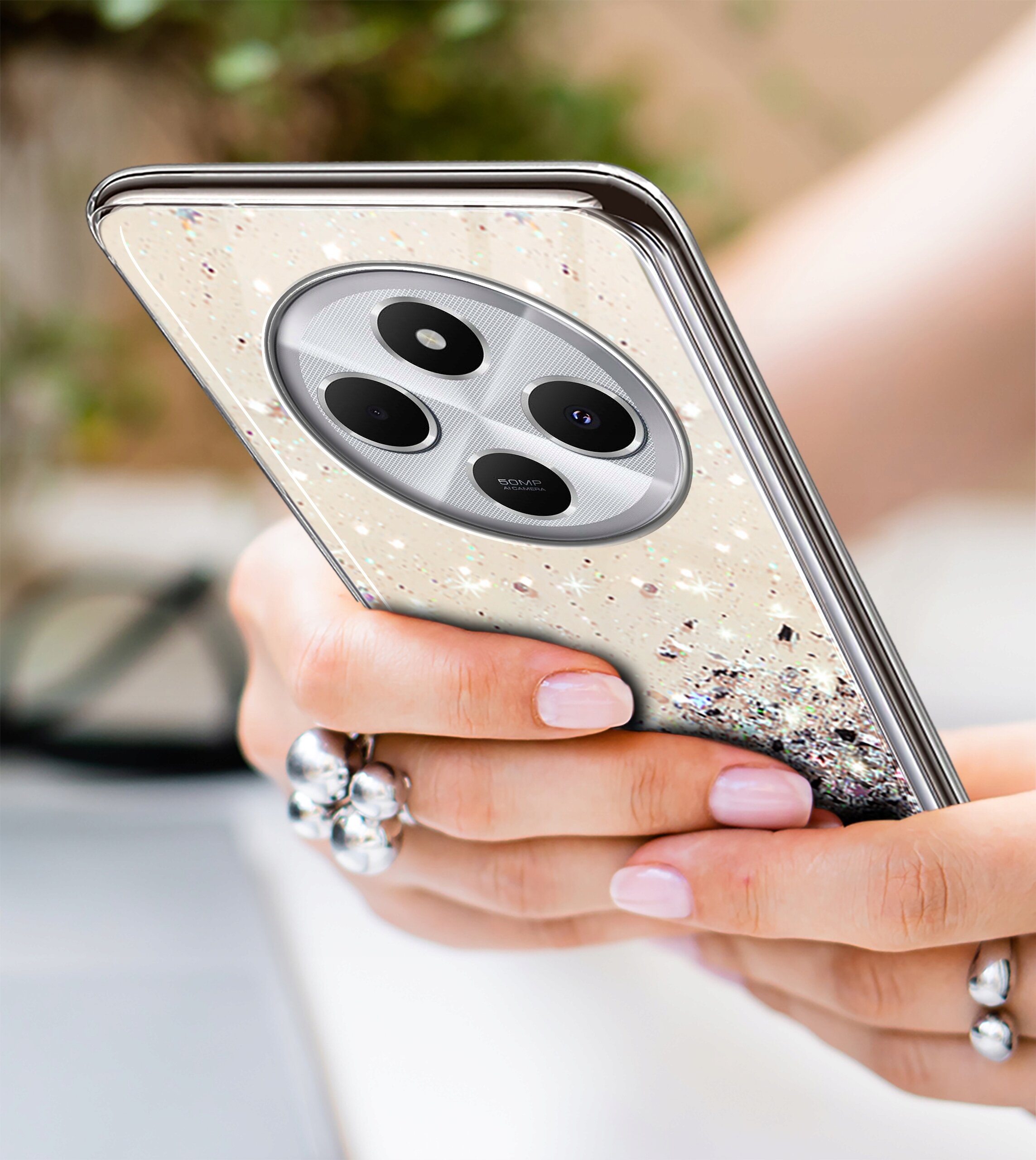 Osoba trzyma telefon w etui z brokatowym wzorem, widoczny jest aparat z trzema obiektywami. W tle znajduje się rozmyty element, prawdopodobnie laptop.