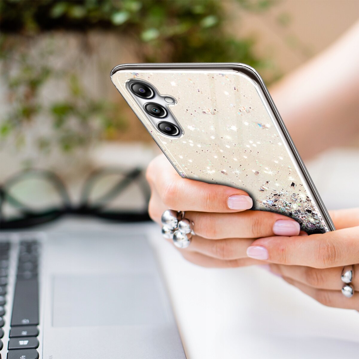 Osoba trzyma telefon z etui brokatowym, w tle widoczny jest fragment laptopa i okulary.