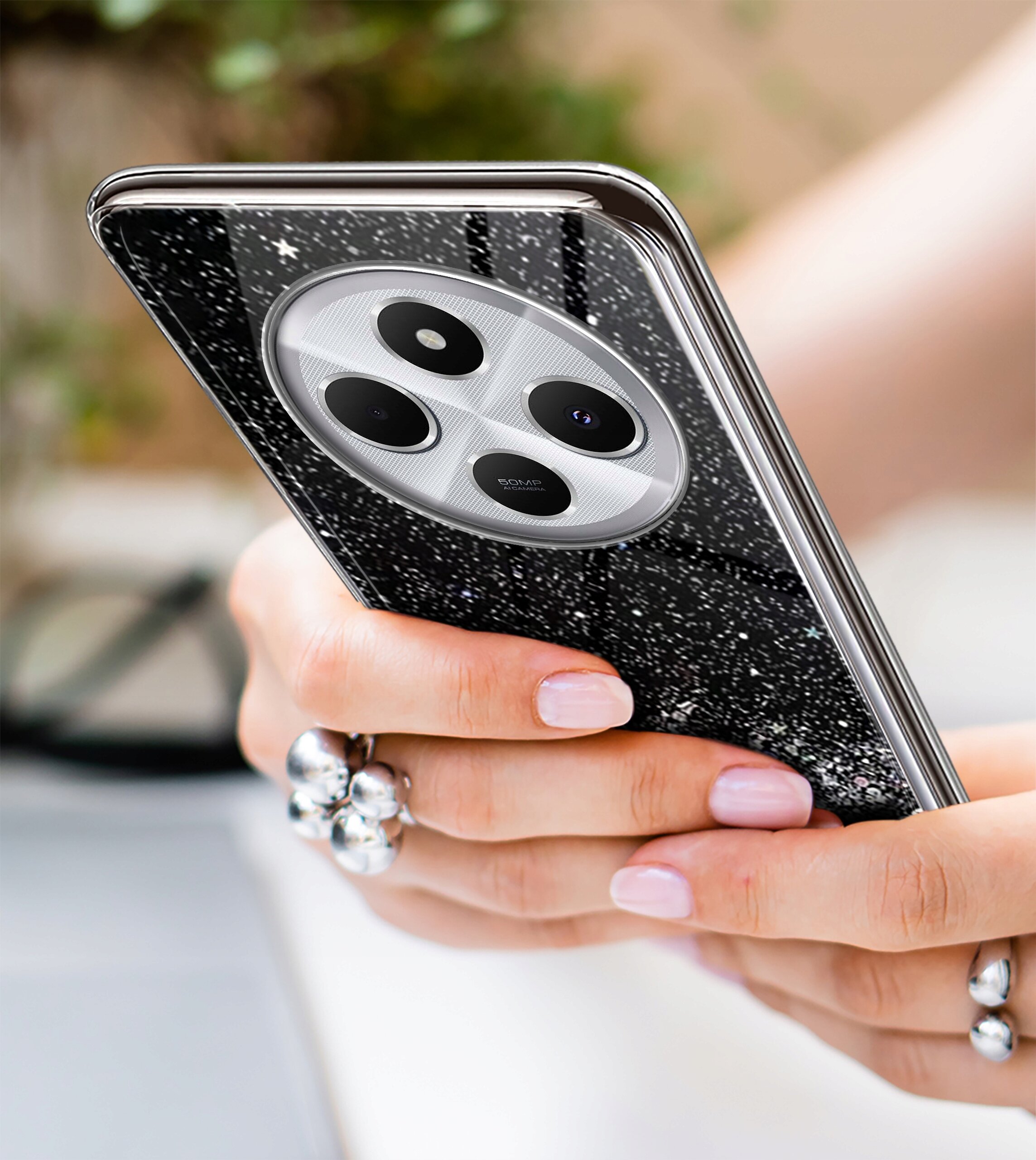 Osoba trzyma telefon w etui z brokatowym wzorem, widoczny jest aparat z trzema obiektywami. W tle znajduje się rozmyte otoczenie z zielenią.
