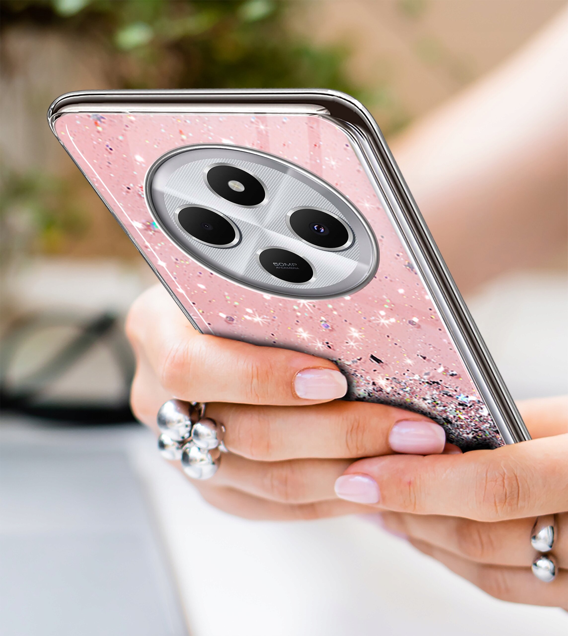 Osoba trzyma telefon w różowym etui z brokatem, widoczny jest aparat z trzema obiektywami. W tle rozmyte elementy, a na palcu pierścionek z dużymi ozdobami.