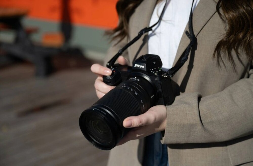 Osoba trzyma aparat fotograficzny z obiektywem marki Nikon. Na aparacie widoczne jest logo \'Nikon\'.