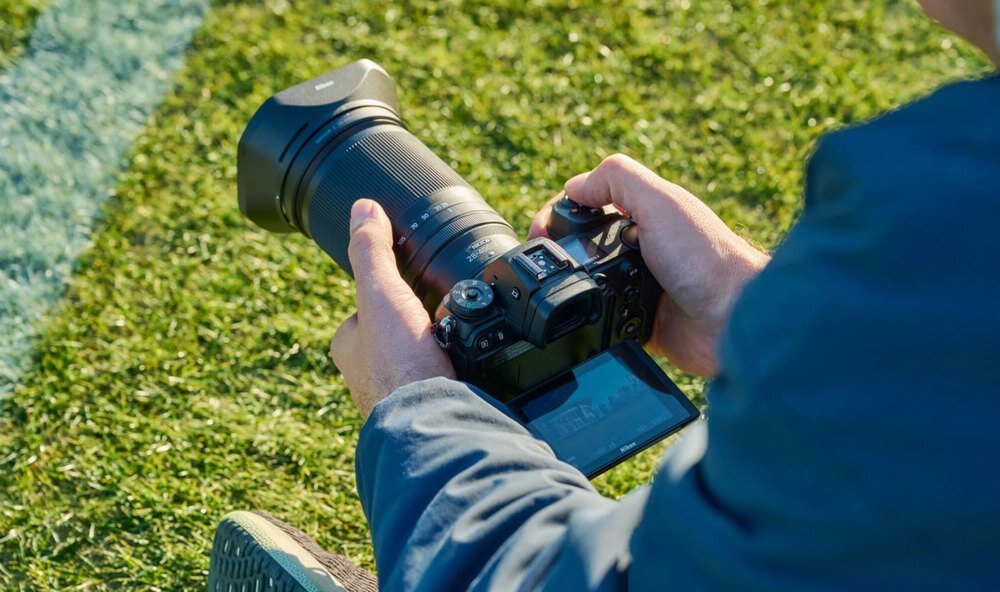 Osoba trzyma aparat fotograficzny z dużym obiektywem, siedząc na trawie. Ekran aparatu jest widoczny, a obiektyw jest skierowany w stronę otoczenia.
