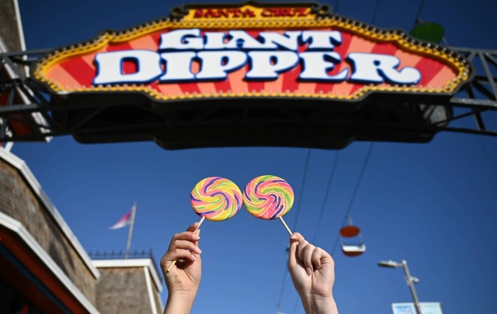Dwie ręce trzymają kolorowe lizaki na tle szyldu z napisem \'GIANT DIPPER\'. W tle widać niebo i fragmenty konstrukcji.
