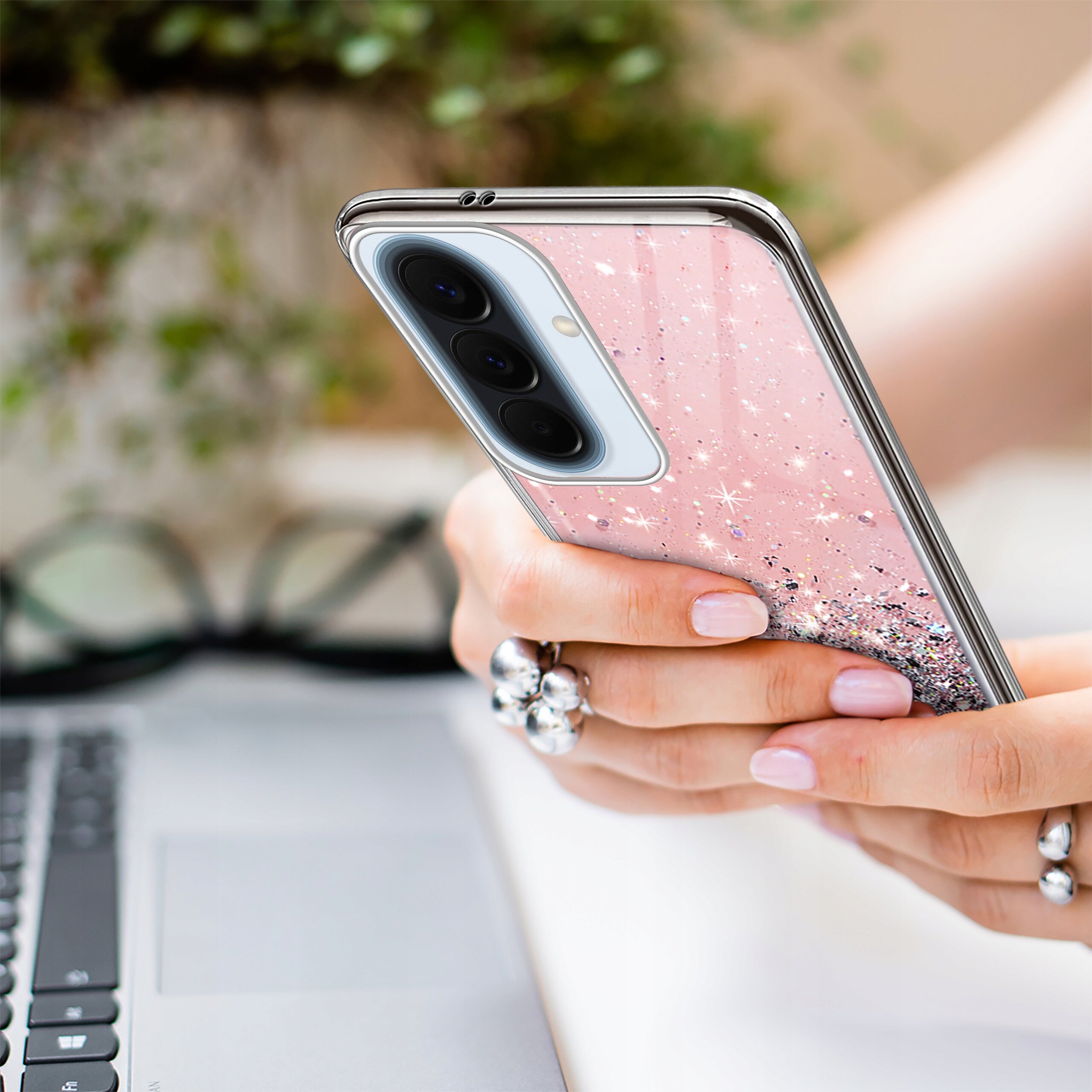 Osoba trzyma telefon w różowym, brokatowym etui, a w tle widoczna jest klawiatura laptopa i okulary.