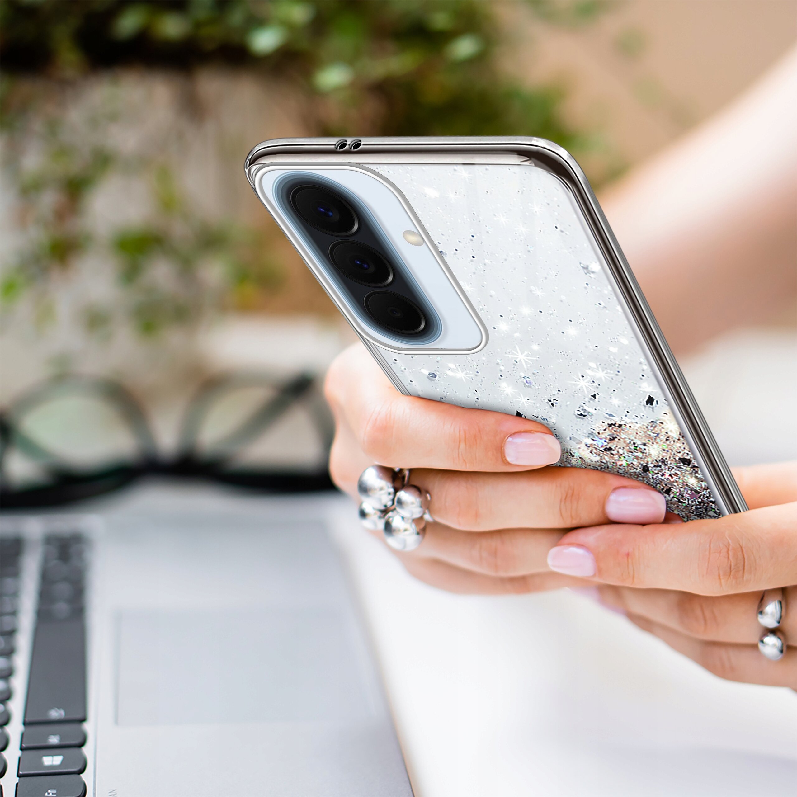 Osoba trzyma telefon z etui brokatowym, a w tle widoczna jest klawiatura laptopa i okulary.