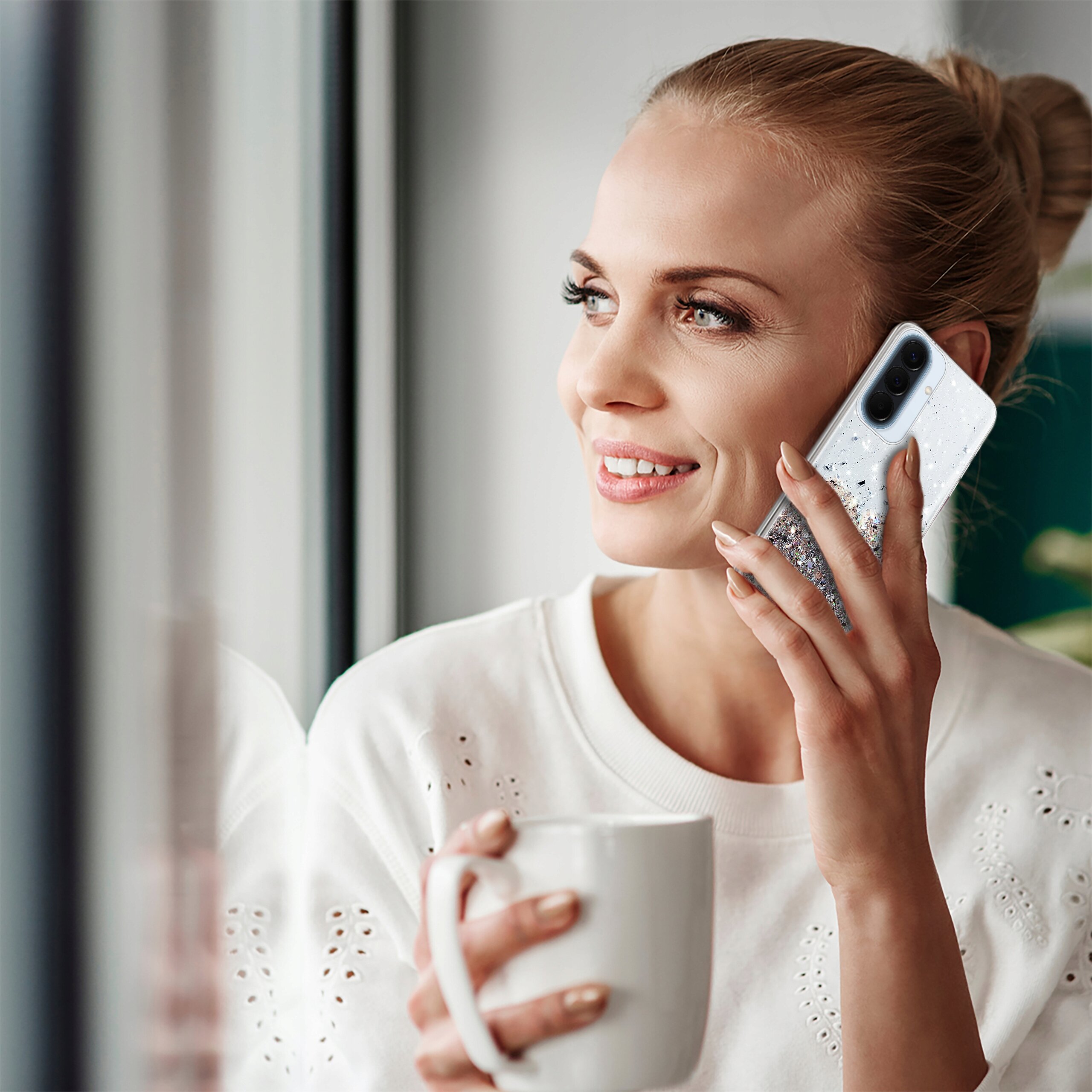 Osoba trzyma telefon z etui brokatowym przy uchu i filiżankę w dłoni. Etui ma błyszczący wzór i przezroczystą część na aparat.