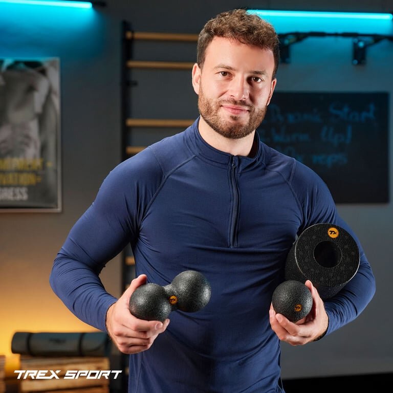 Osoba trzyma zestaw do masażu składający się z czarnego wałka, piłki i duoball z logo TREX SPORT. W tle widoczne są elementy wyposażenia siłowni oraz plakat.