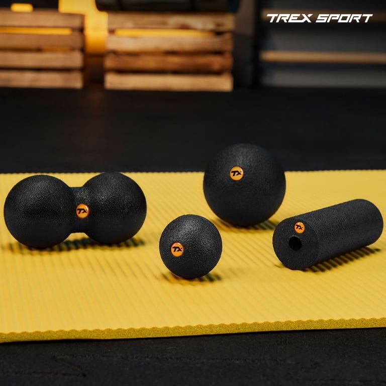 Na żółtej macie leżą cztery czarne akcesoria do masażu: wałek, piłka, duoball i mini roller, każdy z pomarańczowym logo. W tle widoczne są drewniane skrzynki, a w prawym górnym rogu napis \'TREX SPORT\'.