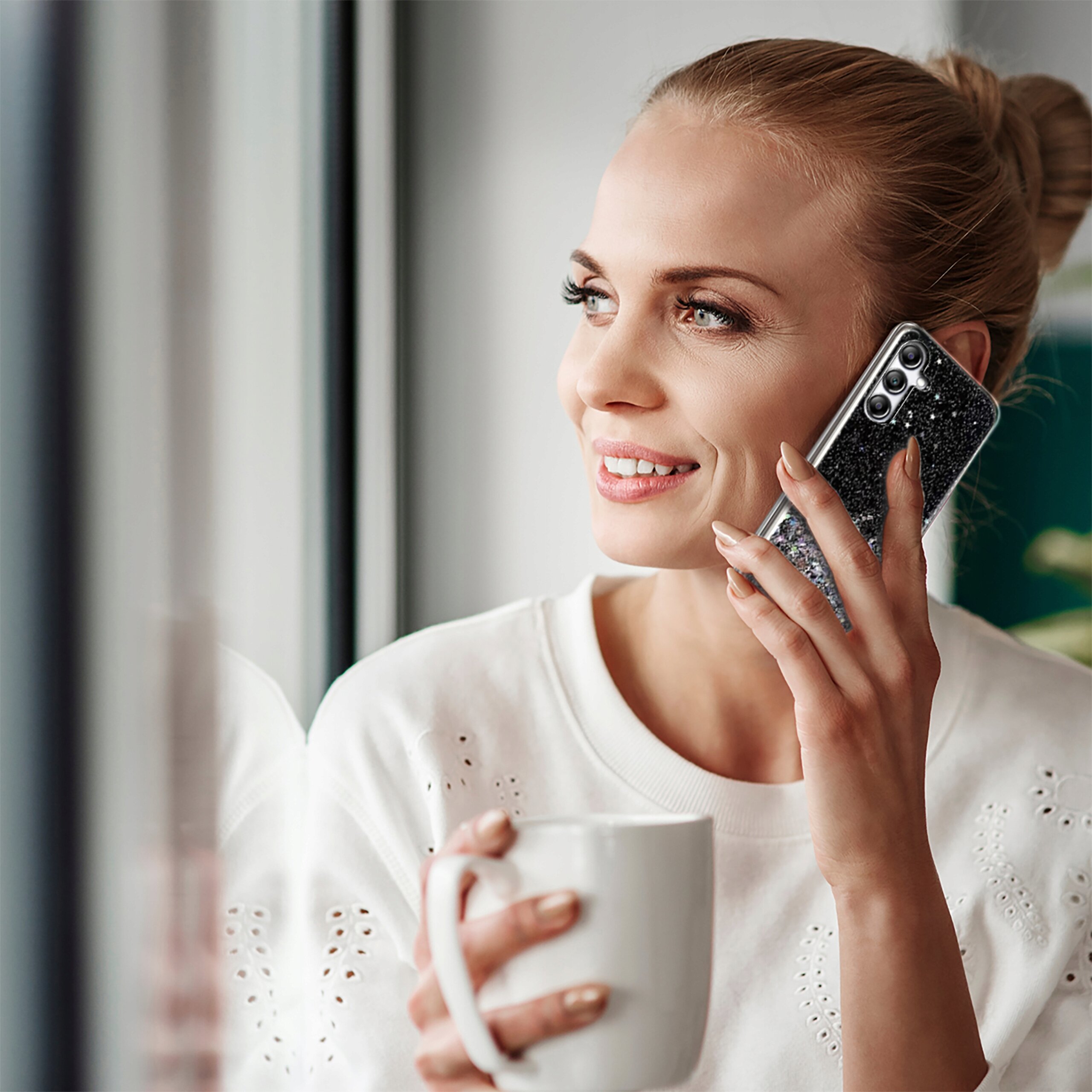 Osoba trzymająca telefon z etui brokatowym przy uchu, w drugiej ręce trzyma kubek.
