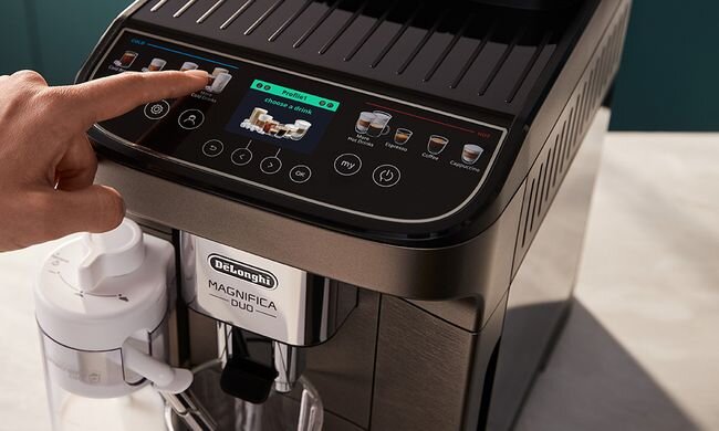 Ekspres do kawy z wyświetlaczem dotykowym, na którym widoczny jest napis \'choose a drink\'. Na obudowie znajduje się logo \'DeLonghi Magnifica Duo\'.