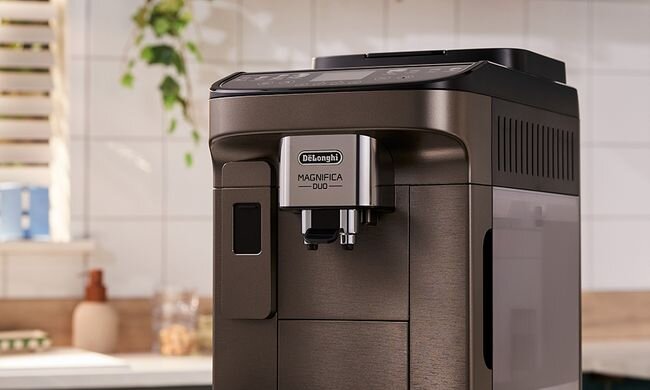 Na obrazie widoczny jest ekspres do kawy w kolorze ciemnym, z logo \'DeLonghi\' oraz napisem \'MAGNIFICA DUO\' na przednim panelu. W tle znajduje się kuchnia z białymi kafelkami i rośliną.