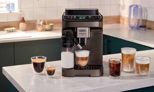Na blacie kuchennym stoi ekspres do kawy z logo \'DeLonghi Magnifica S\'. Obok ekspresu znajduje się kilka szklanek z różnymi napojami kawowymi.