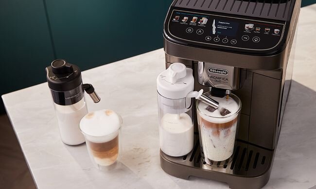 Na blacie stoi ekspres do kawy z logo \'DeLonghi Magnifica Duo\', obok znajdują się dwa kubki z kawą i dwa pojemniki na mleko.