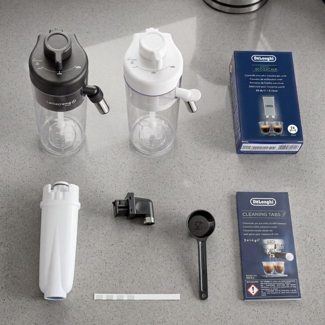 Na blacie znajdują się dwa pojemniki z pokrywkami, jeden czarny, drugi biały, oraz pudełko z napisem \'DeLonghi ECAM\'. Obok leżą różne akcesoria, w tym filtr, łyżeczka i opakowanie \'DeLonghi CLEANING TABS\'.