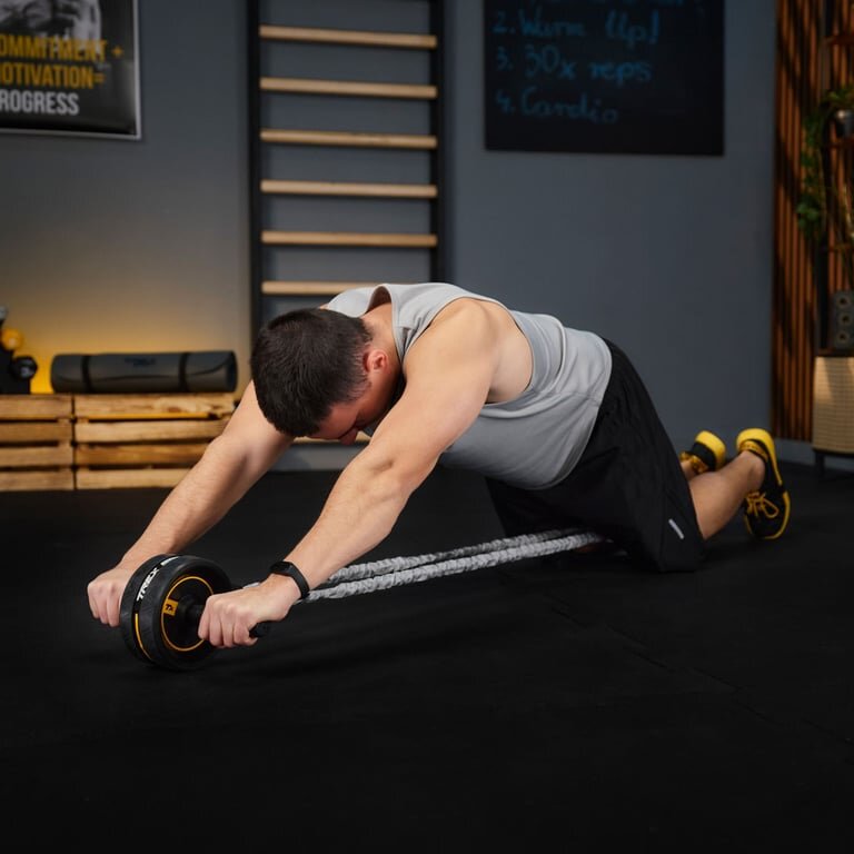 Osoba ćwiczy na macie, używając kółka do mięśni brzucha. W tle widoczne są drewniane palety i tablica z napisami: 2. Warm (up!), 3. 30x reps, 4. Cardio.