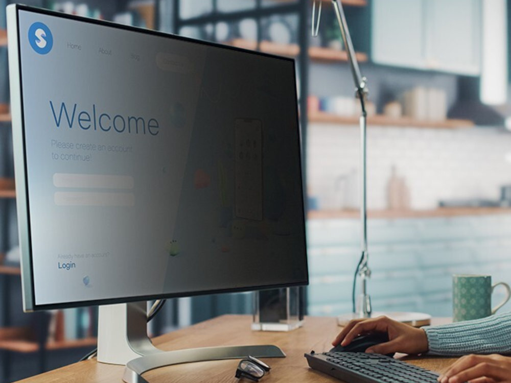 Na biurku stoi monitor z widocznym ekranem, na którym wyświetlany jest tekst: \'Welcome Please create an account to continue! Login\'. W tle znajduje się rozmyta kuchnia, a na biurku leży klawiatura i kubek.