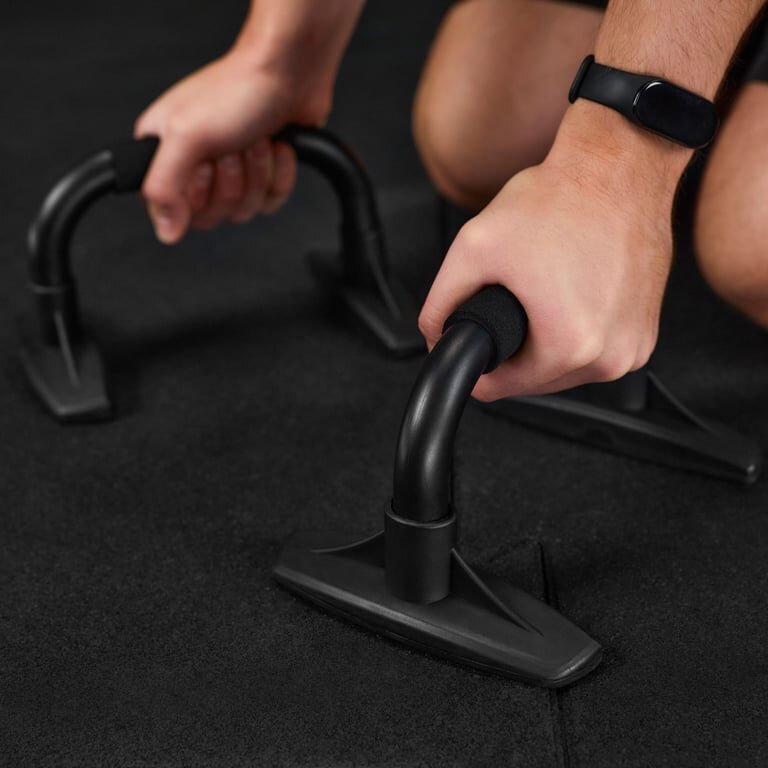 Osoba trzyma czarne uchwyty do pompek na ciemnej macie do ćwiczeń. Na nadgarstku widoczna jest opaska fitness.