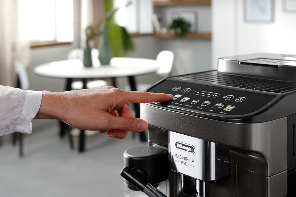 Osoba dotyka panelu sterowania ekspresu do kawy z widocznymi ikonami napojów. Na urządzeniu znajduje się logo \'DeLonghi\' oraz napis \'MAGNIFICA EVO\'.