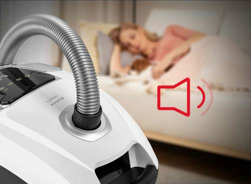 Na pierwszym planie widoczny jest biały odkurzacz z napisem \'Amica Suracon Pet VM 7003\', a w tle osoba leży na łóżku z psem. Obok odkurzacza znajduje się czerwony symbol głośności.