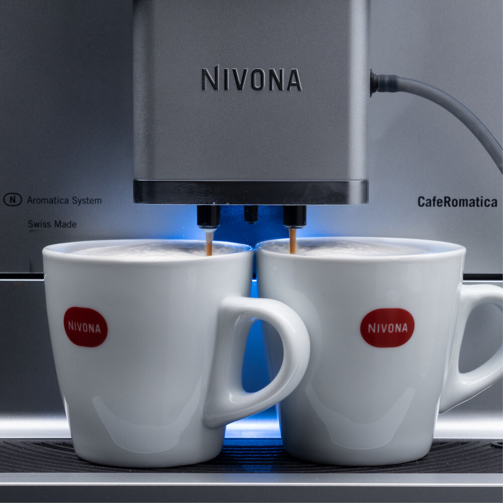 Ekspres do kawy z logo NIVONA przygotowuje kawę do dwóch białych filiżanek z napisem NIVONA. Na urządzeniu widoczne są napisy: Aromatica System, Swiss Made, CafeRomatica.