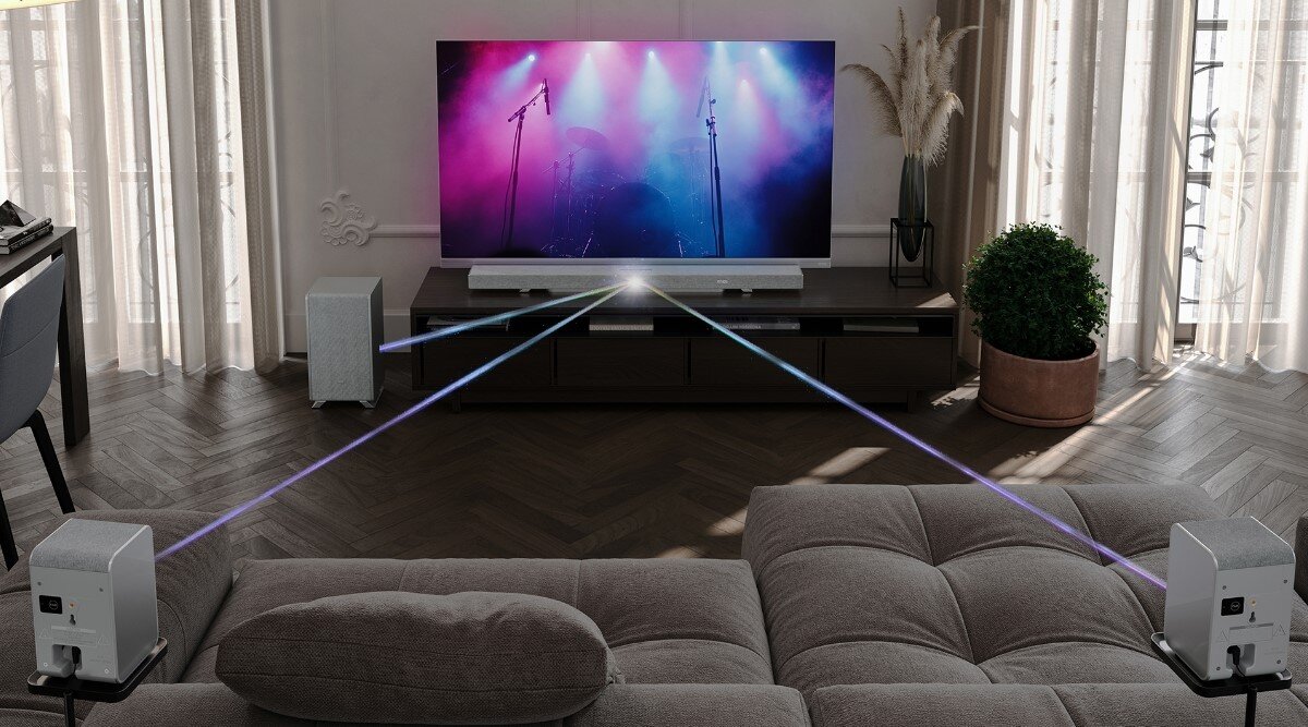 W salonie znajduje się telewizor z wyświetlanym kolorowym obrazem sceny koncertowej, przed którym ustawiony jest soundbar. Wokół rozstawione są głośniki, a na podłodze leży dywan i stoi roślina w doniczce.