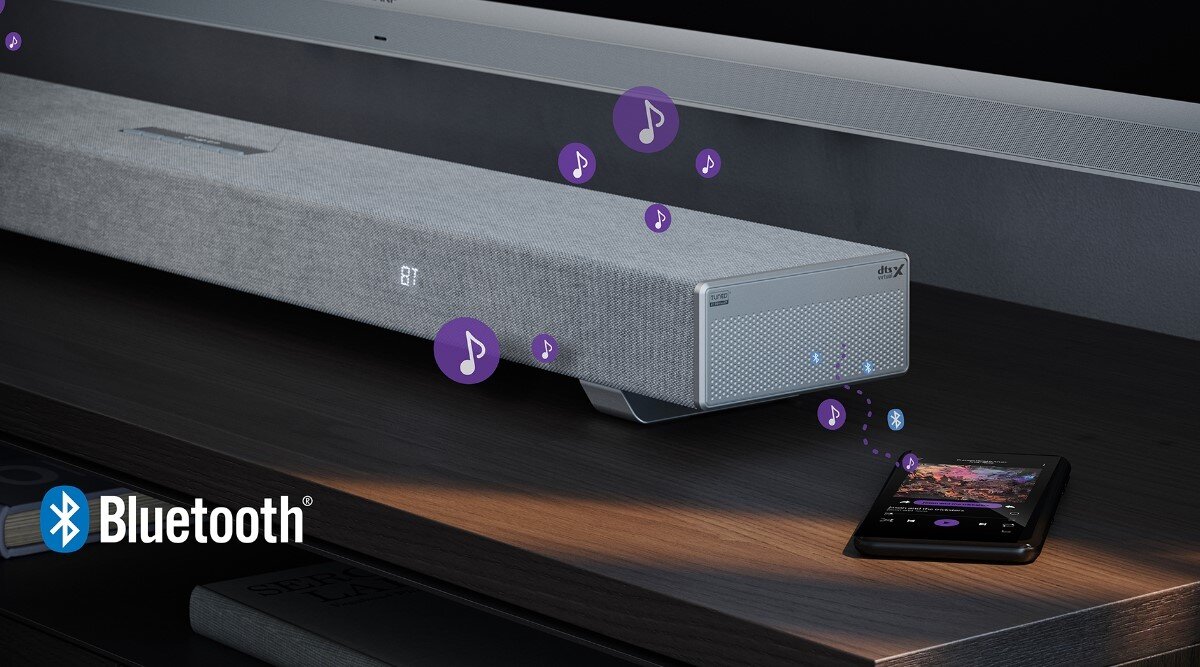 Na obrazie widoczny jest srebrny soundbar z logo \'dtsX\' na froncie, umieszczony na drewnianej powierzchni. Obok znajduje się smartfon, z którego unoszą się ikony nutek, a w lewym dolnym rogu widnieje logo \'Bluetooth\'.
