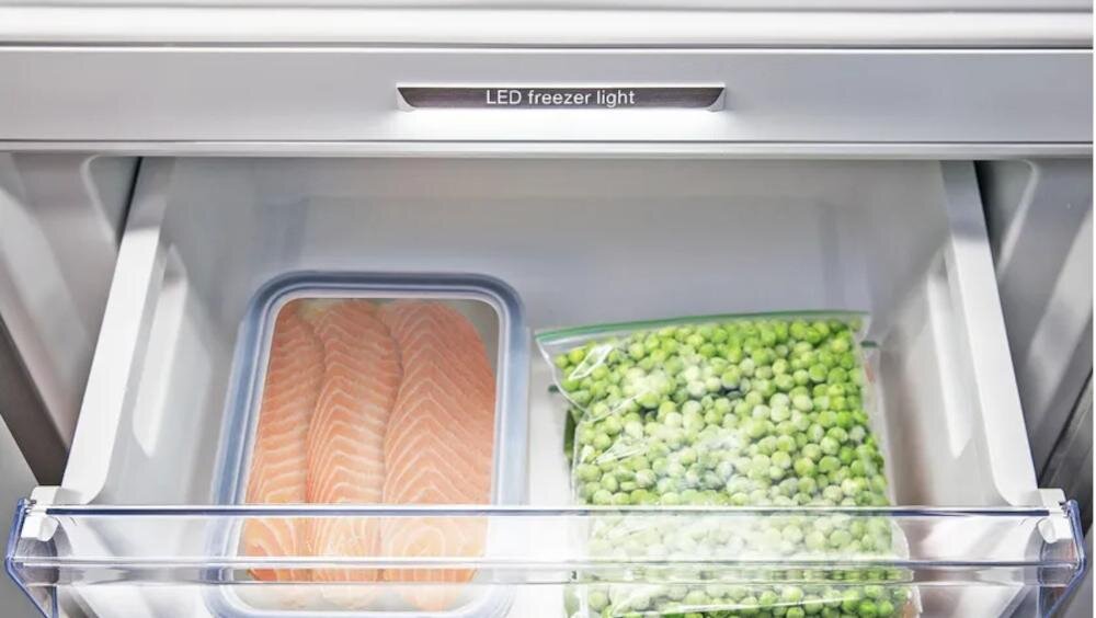 Wnętrze szuflady zamrażarki z oświetleniem LED, zawierające pojemnik z plastrami łososia oraz worek z zielonym groszkiem. Widoczny napis: \'LED freezer light\'.