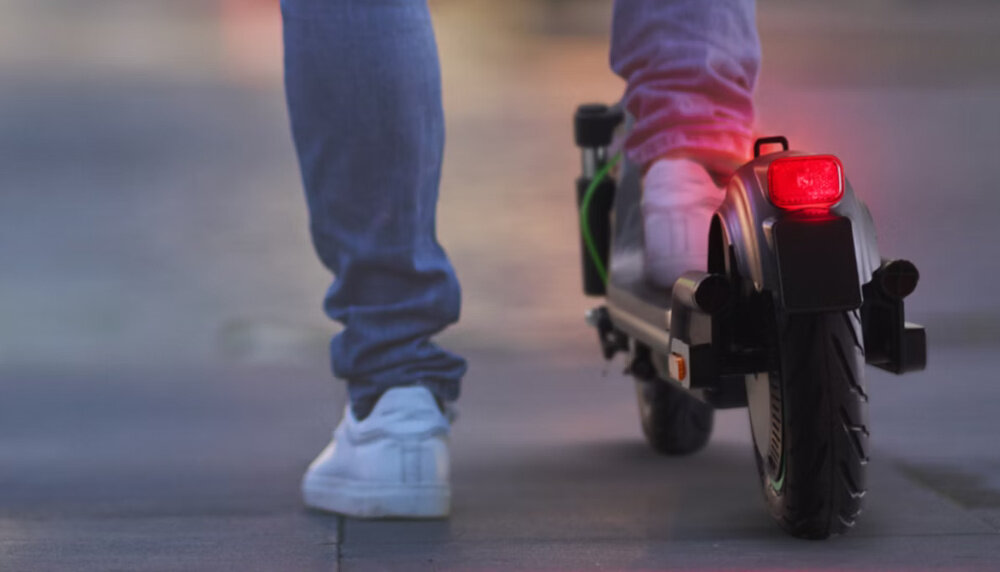 Osoba w białych butach stoi na elektrycznej hulajnodze z włączonym tylnym światłem. Hulajnoga ma czarny kolor i widoczne są jej tylne koło oraz elementy amortyzacji.
