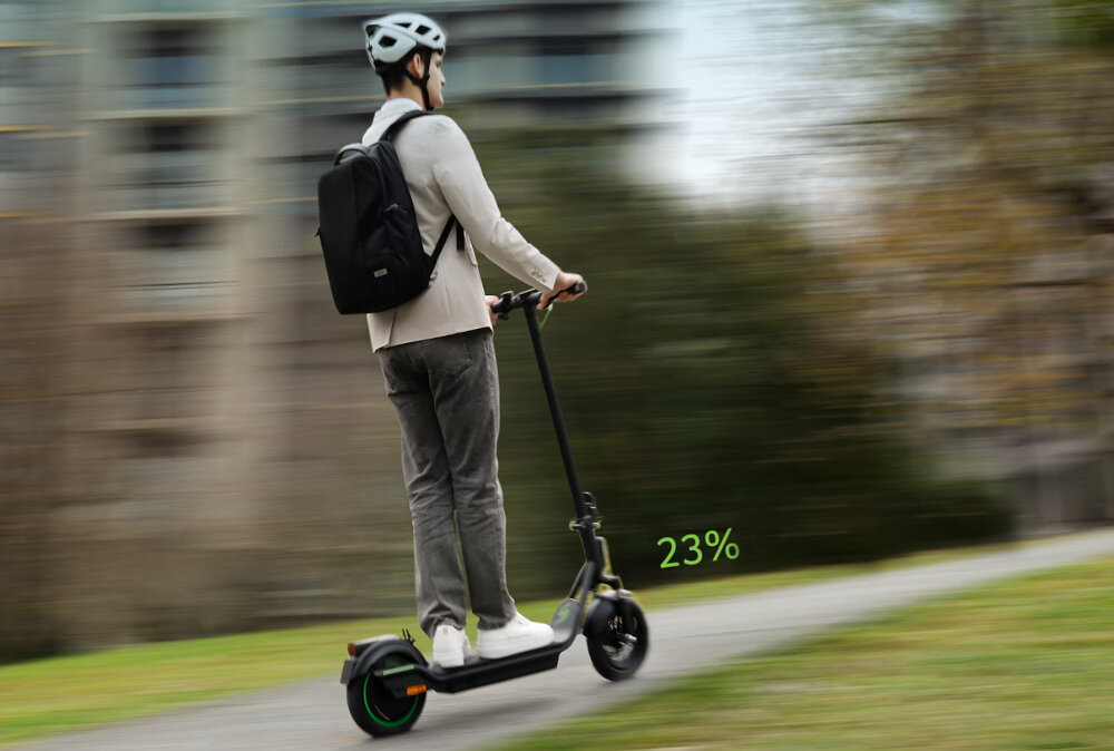 Osoba w kasku jedzie na czarnej hulajnodze elektrycznej po ścieżce w parku. Na obrazie widoczny jest napis \'23%\'.