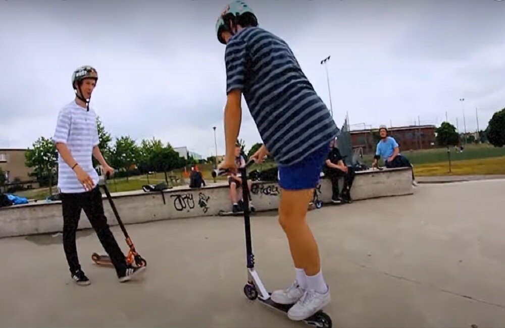 Dwóch młodych ludzi na skateparku, każdy z nich trzyma hulajnogę wyczynową. W tle widoczne są graffiti na murku oraz kilka osób siedzących na ławce.