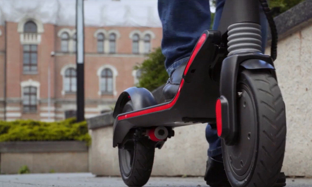 Osoba stoi na czarnej hulajnodze elektrycznej z czerwonymi akcentami, widoczny jest tylny amortyzator i koło. W tle znajduje się budynek z ozdobnymi oknami.