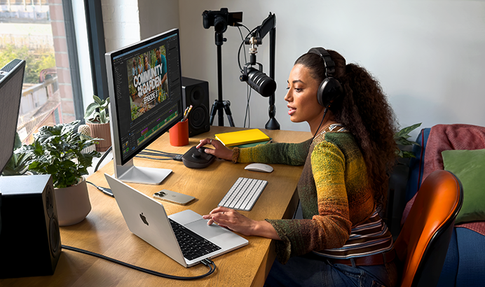 Osoba siedzi przy biurku z laptopem marki APPLE, monitorem, klawiaturą i mikrofonem. Na ekranie monitora widoczny jest tekst \'COMMUNITY GARDEN\'.