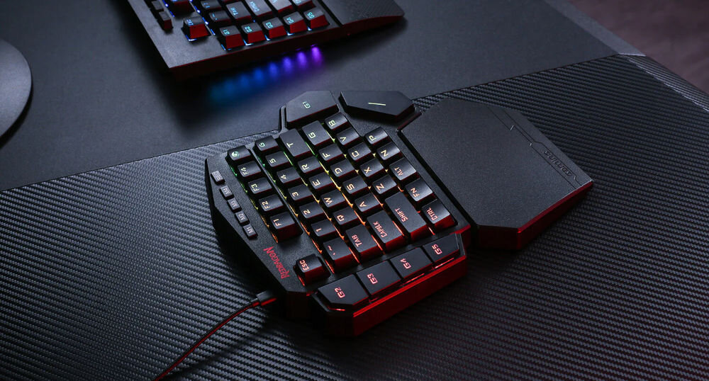 Na obrazie widoczna jest klawiatura gamingowa z podświetleniem RGB, umieszczona na ciemnym biurku o fakturze przypominającej włókno węglowe. Na klawiaturze znajduje się logo \'Redragon\'.
