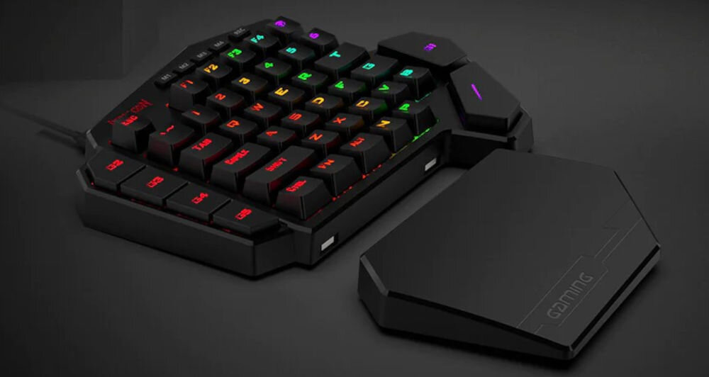 Na obrazie widoczna jest klawiatura gamingowa z podświetleniem RGB, posiadająca kilka rzędów klawiszy. Obok klawiatury znajduje się odłączana podpórka na nadgarstek z napisem \'GAMING\'.