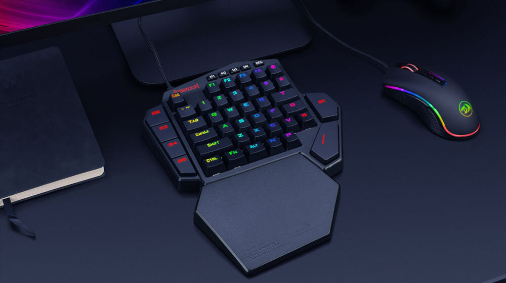 Na ciemnym biurku znajduje się klawiatura z podświetlanymi klawiszami RGB oraz mysz z podświetleniem. Obok leży zamknięty notes i widoczny jest kabel klawiatury.