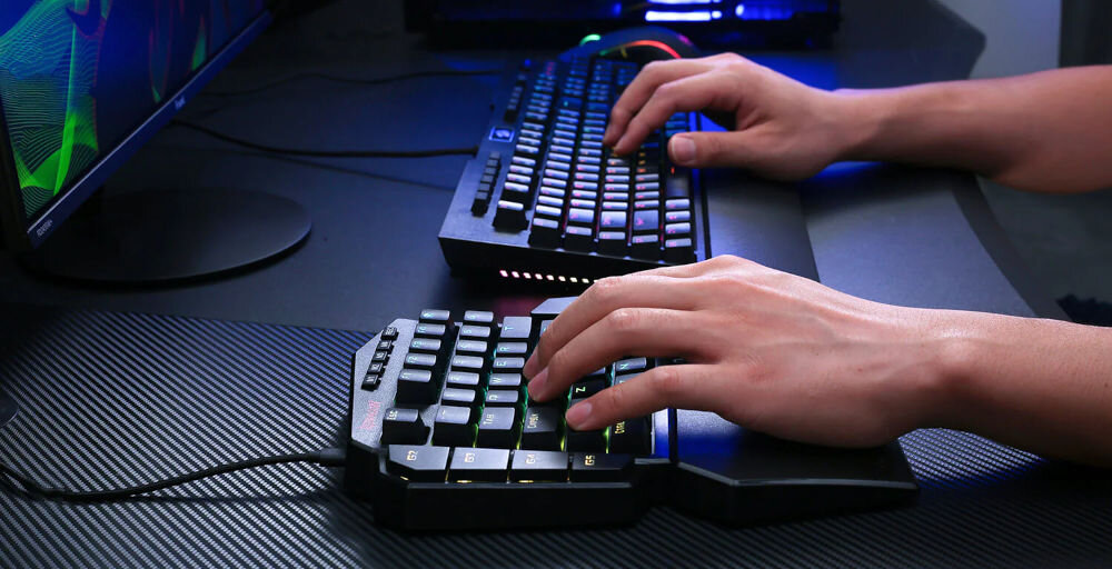 Osoba używa klawiatury z podświetleniem RGB, widoczna jest także druga klawiatura i monitor. Na klawiaturze znajduje się logo REDRAGON.