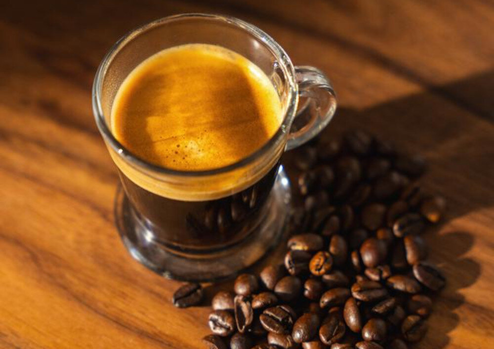 Szklanka espresso z kremową pianką stoi na drewnianym blacie, obok rozsypane są ziarna kawy.