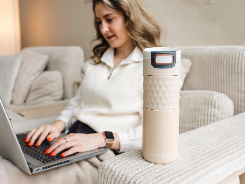 Beżowy kubek termiczny KAMBUKKA Etna Grip 500 ml stoi na kanapie. W tle osoba korzysta z laptopa.