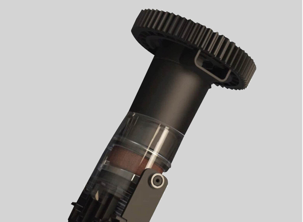 Obraz przedstawia czarny element mechaniczny z przezroczystą częścią, przypominający część ekspresu do kawy, na szarym tle.